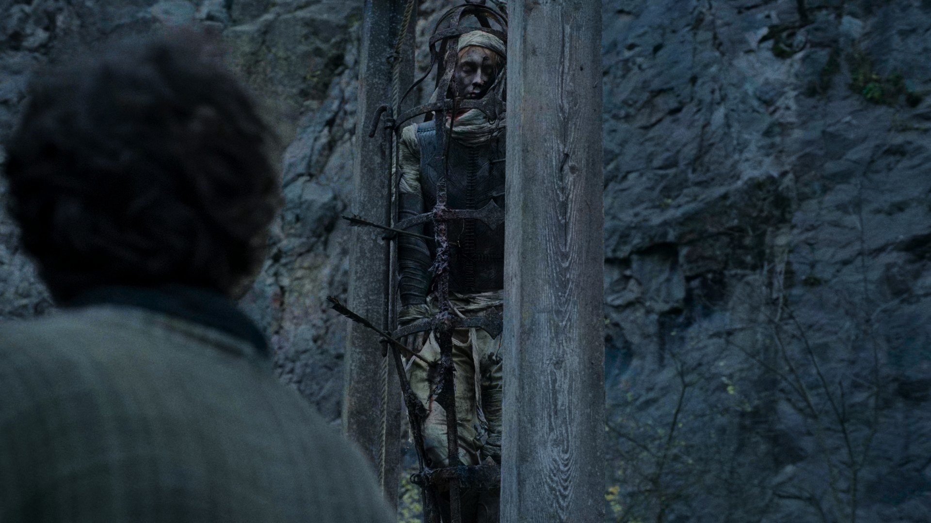 A man in beige clothing and a headscarf is in a cage hanging from a gibbet. He is pierced with many arrows through his torso, which has a stylised leather jerkin on it. He has been dead for a while.