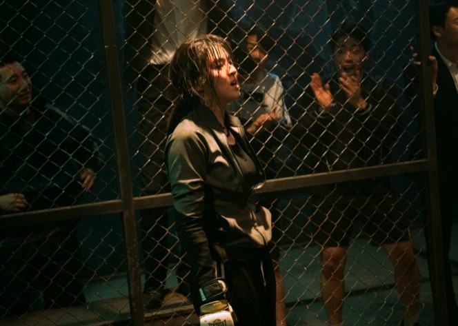 A woman stands in a fighting cage wearing boxing gloves as men cheer in the background