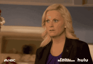 Amy Poehler as Leslie Knope stands in a home and looks on with wide eyes as the camera zooms out to reveal Rashida Jones as Ann Perkins sitting on the couch with an open mouth in