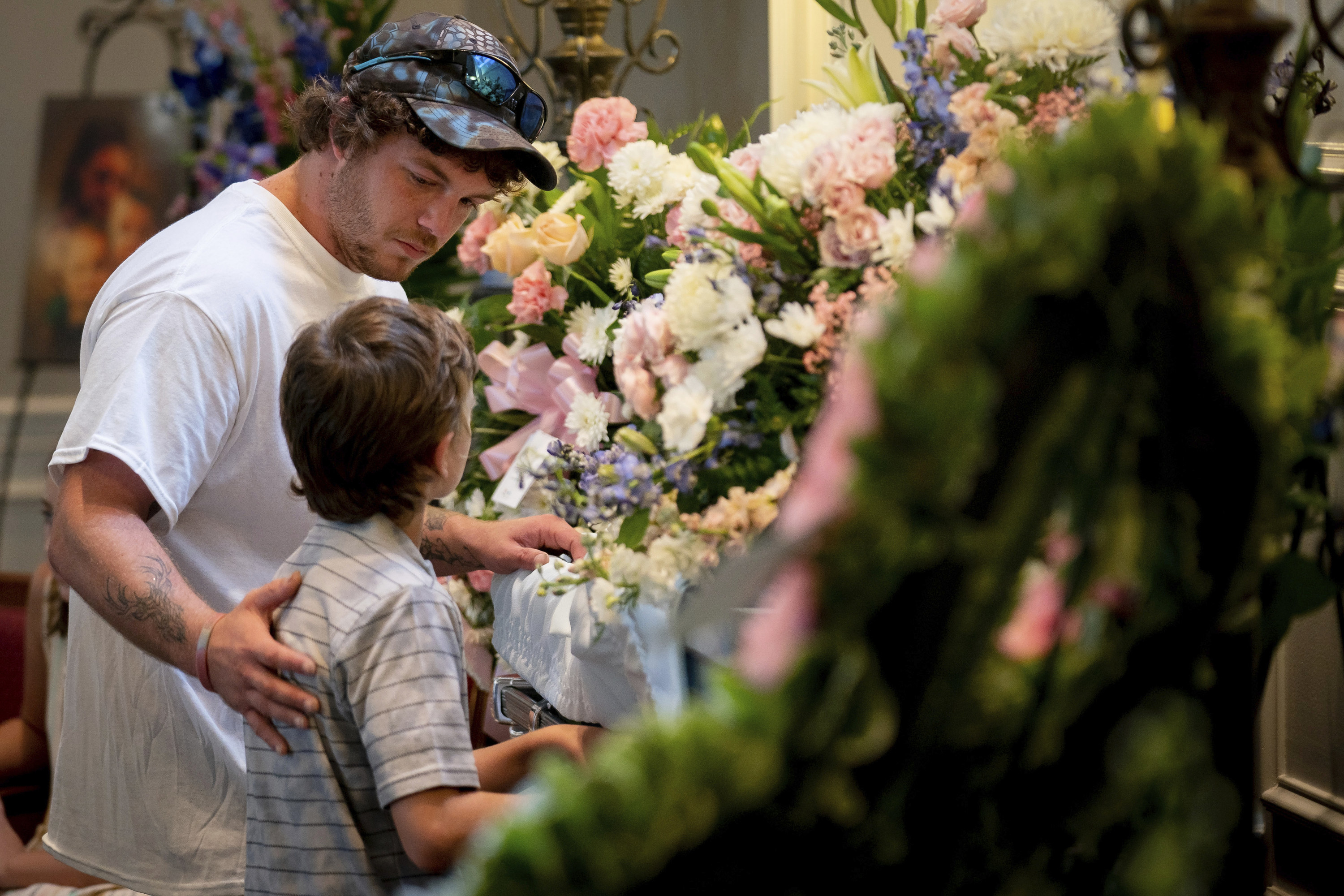 A man and young boy stand in front of a casket covered in flowers