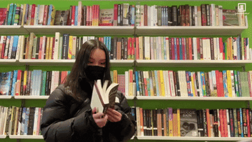 ID: a woman flips through a book in front of a large bookshelf