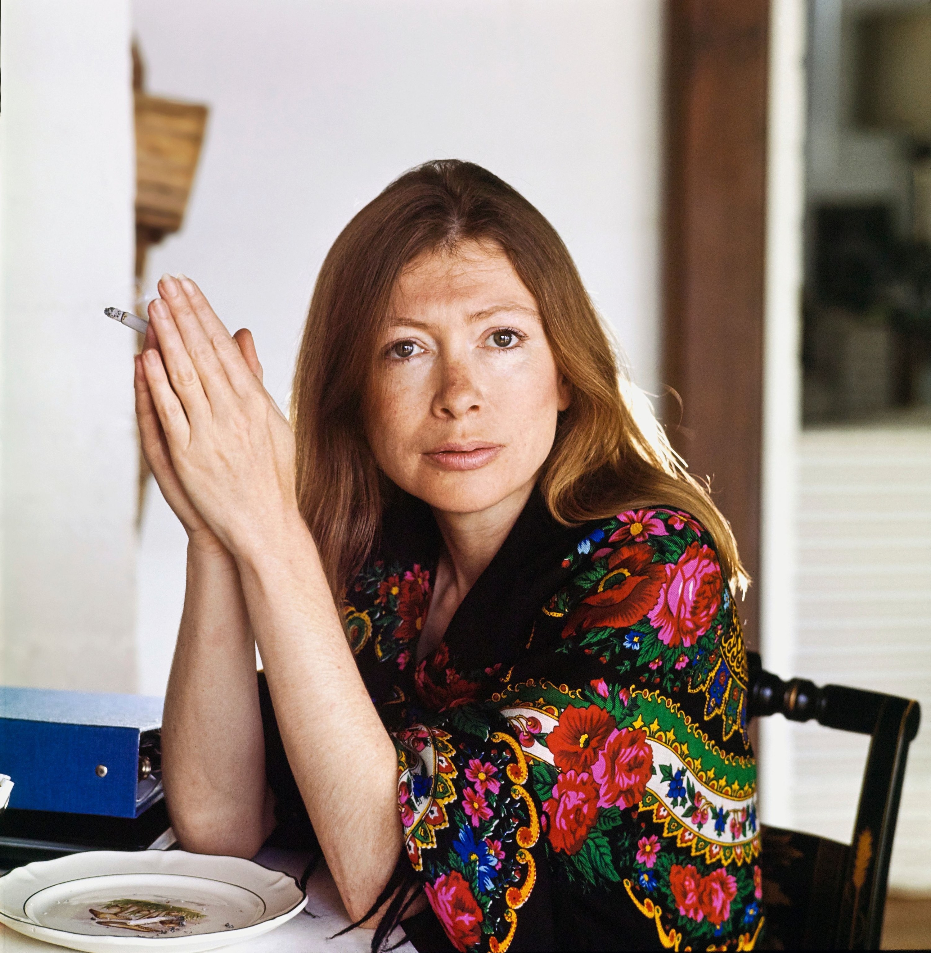 Joan Didion in a robe sitting at a table with her hands pressed together