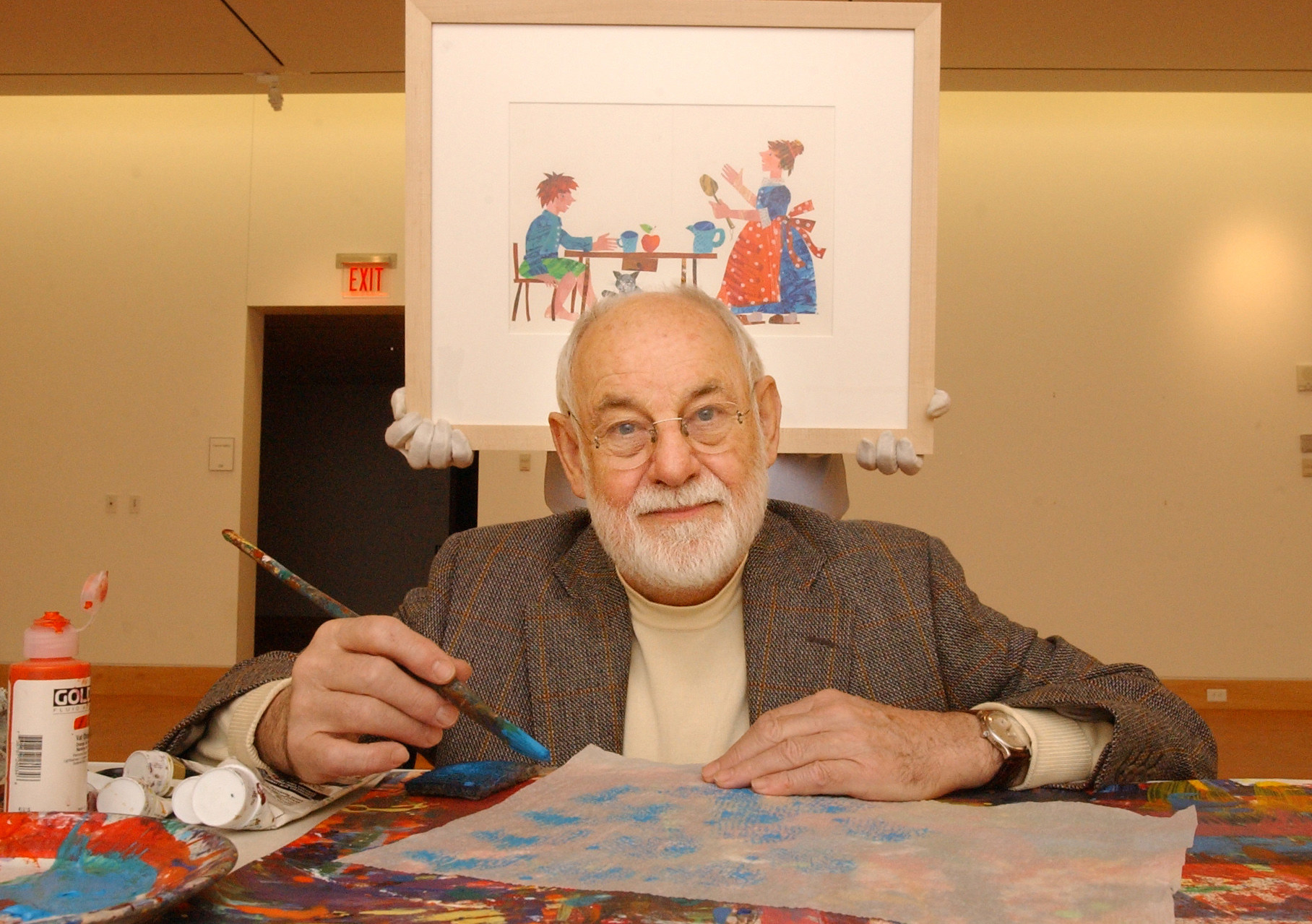 Eric Carle with a paintbrush and paint with someone holding up a framed illustration behind him 