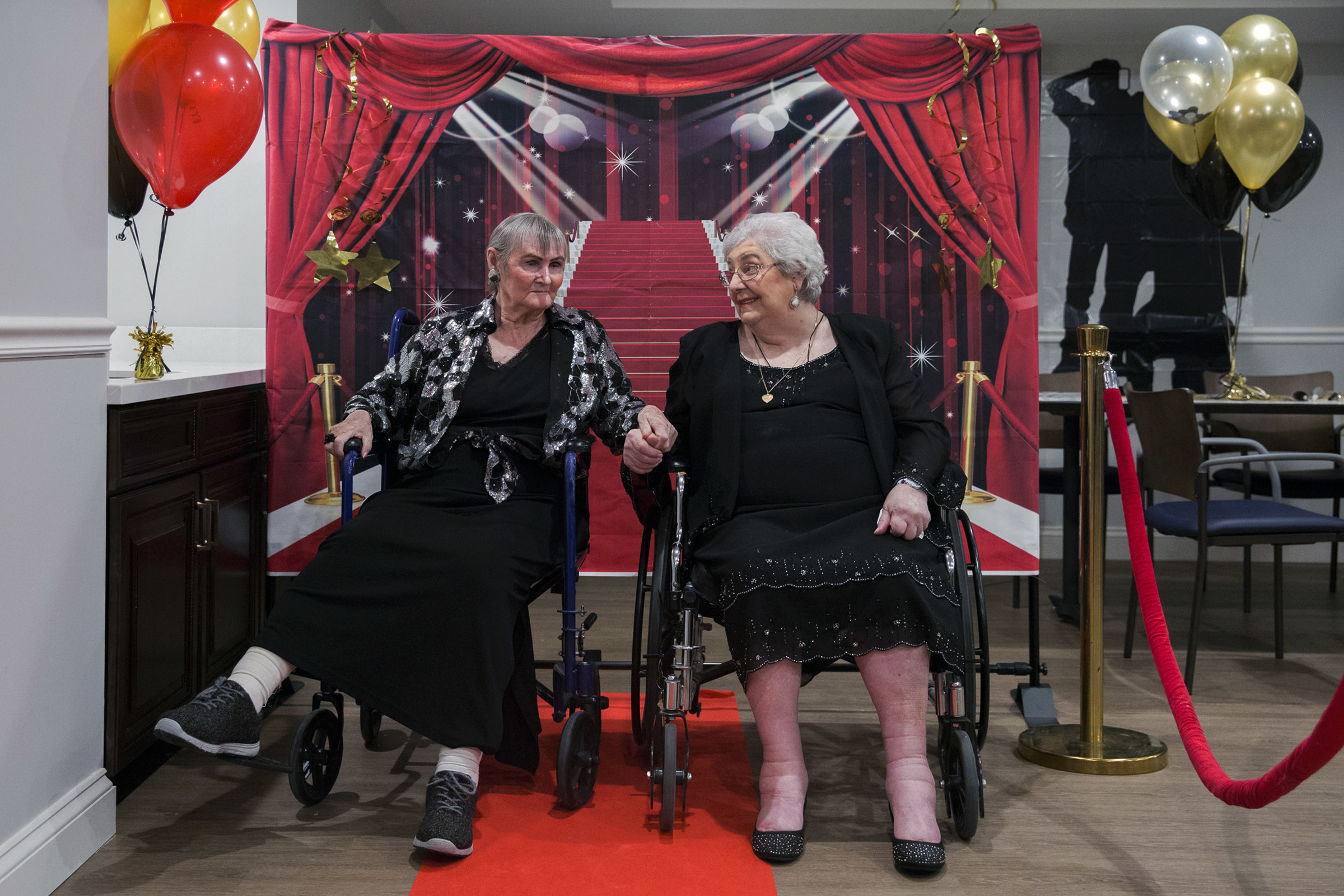 Two older women dressed in black, holding hands while in wheelchairs in front of a red carpet backdrop 