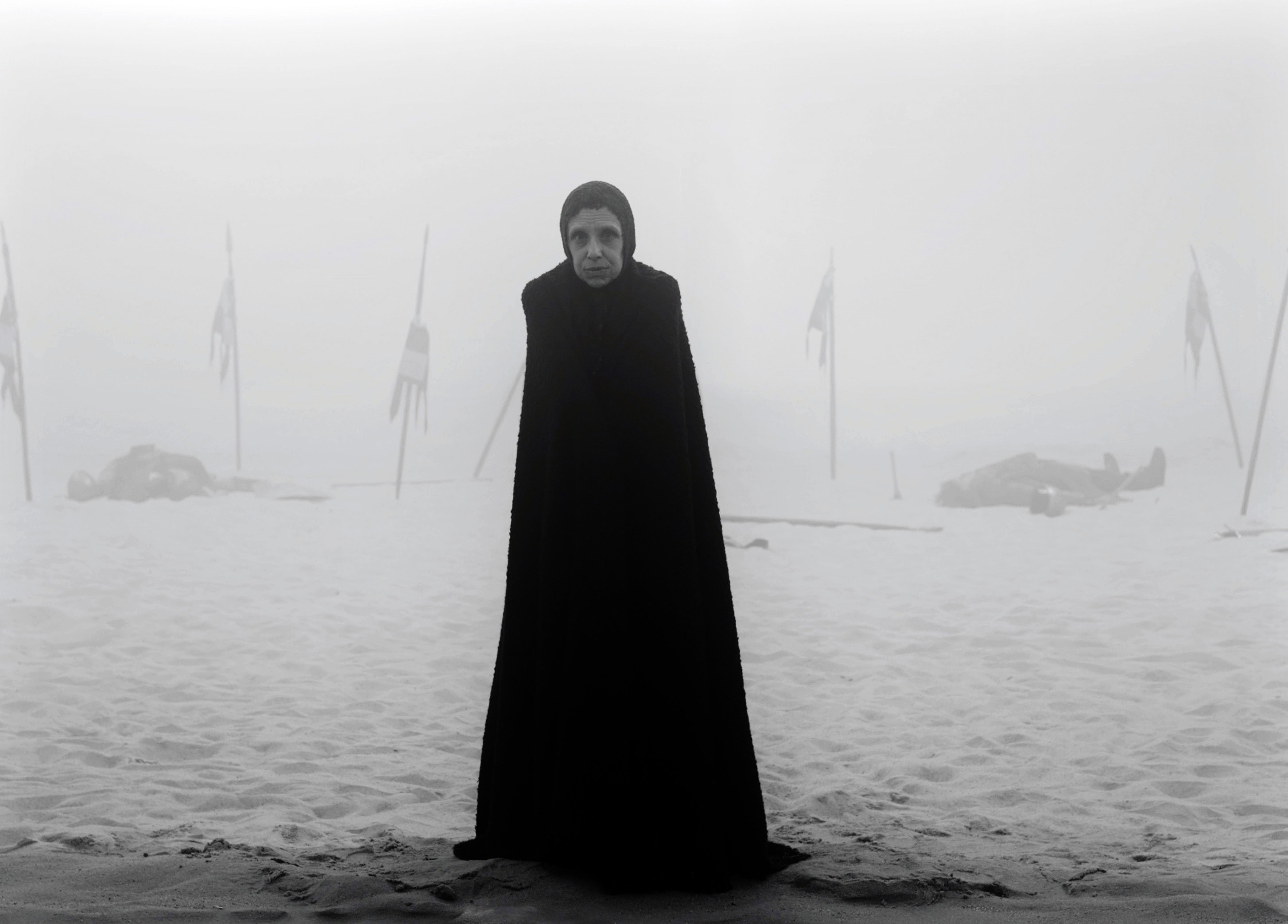 Kathryn Hunter stands on a sandy shore in a cloak
