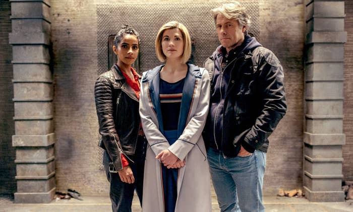 Pro shot shows actors Mandip Gill, Jodie Whittaker, and John Bishop in their Doctor Who roles posing in front of a stone wall