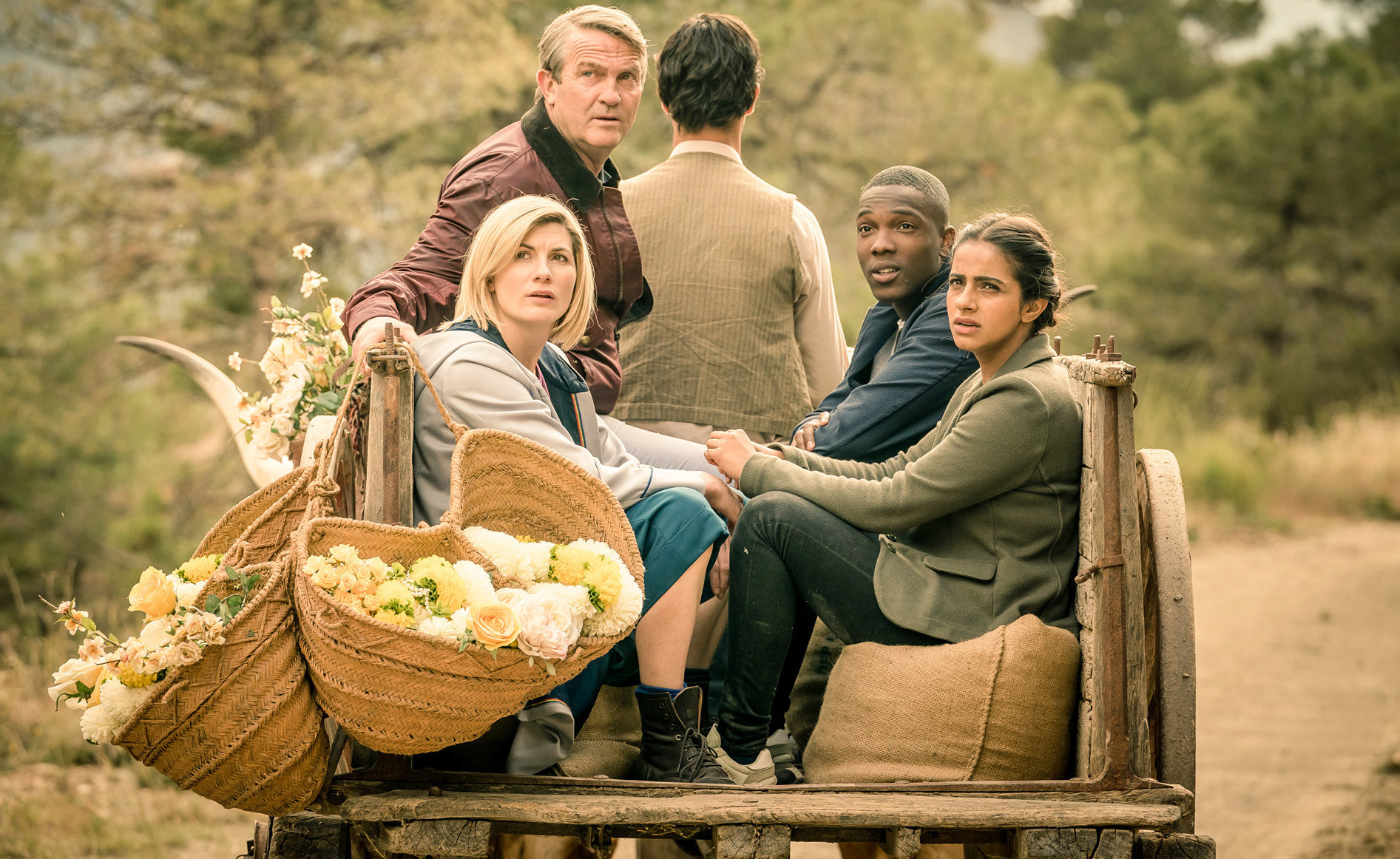 Still from an episode of Doctor Who show the Doctor, Yaz, Ryan, and Graham aboard a cart on a dusty road looking at something off camera