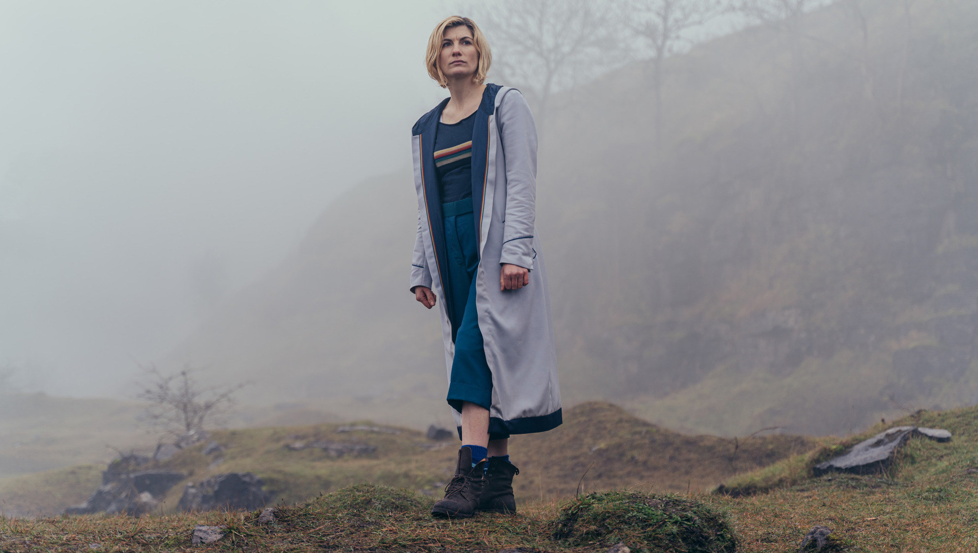 Jodie Whittaker as the Doctor stands on a rock in a long grey coat amongst a foggy rural setting