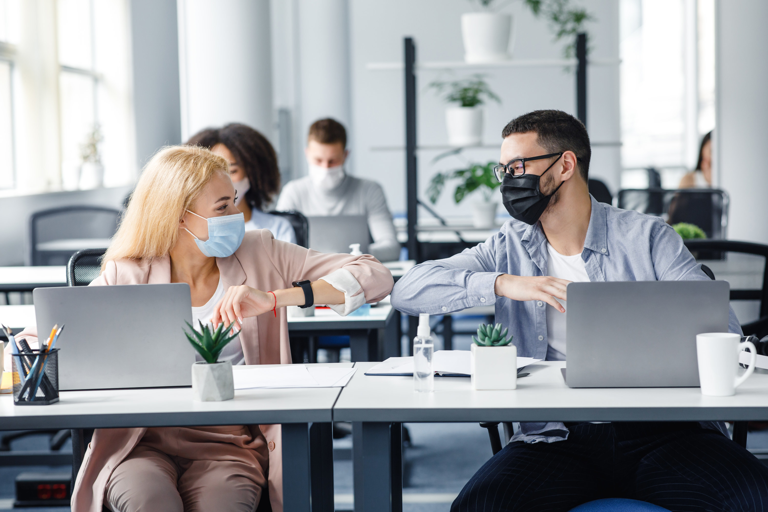 Two co-workers who are sitting in front of their laptops with masks on bumping their elbows