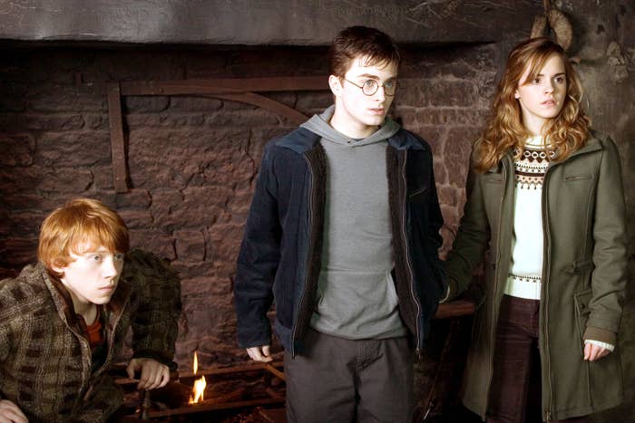 Rupert Grint, Daniel Radcliffe, and Emma Watson stand next to each other in front of a fireplace