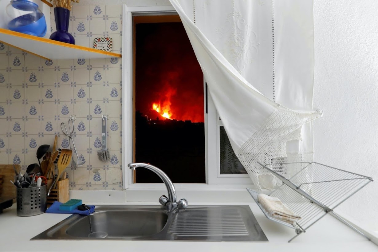 A kitchen with a curtain pulled back, witch a fire raging in the view through the window