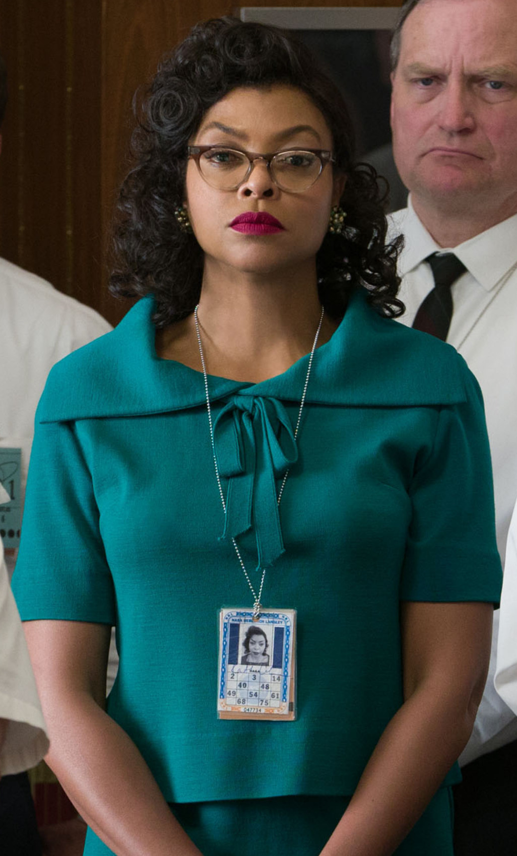 Henson wearing glasses, 1950s dress, and bright lipstick in a meeting surrounded by white men