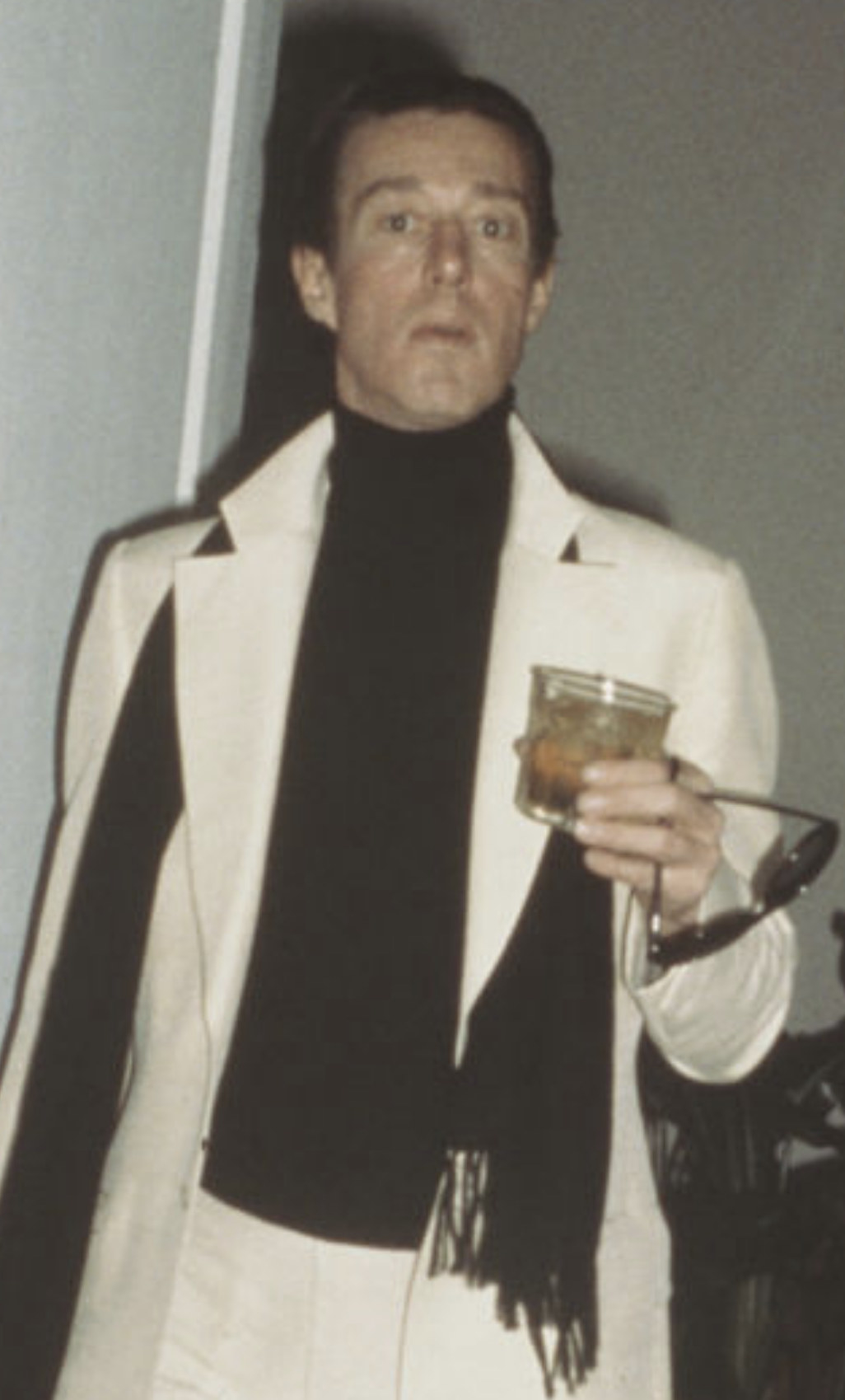 Halston holding a cocktail while wearing a long scarf and matching suit/coat