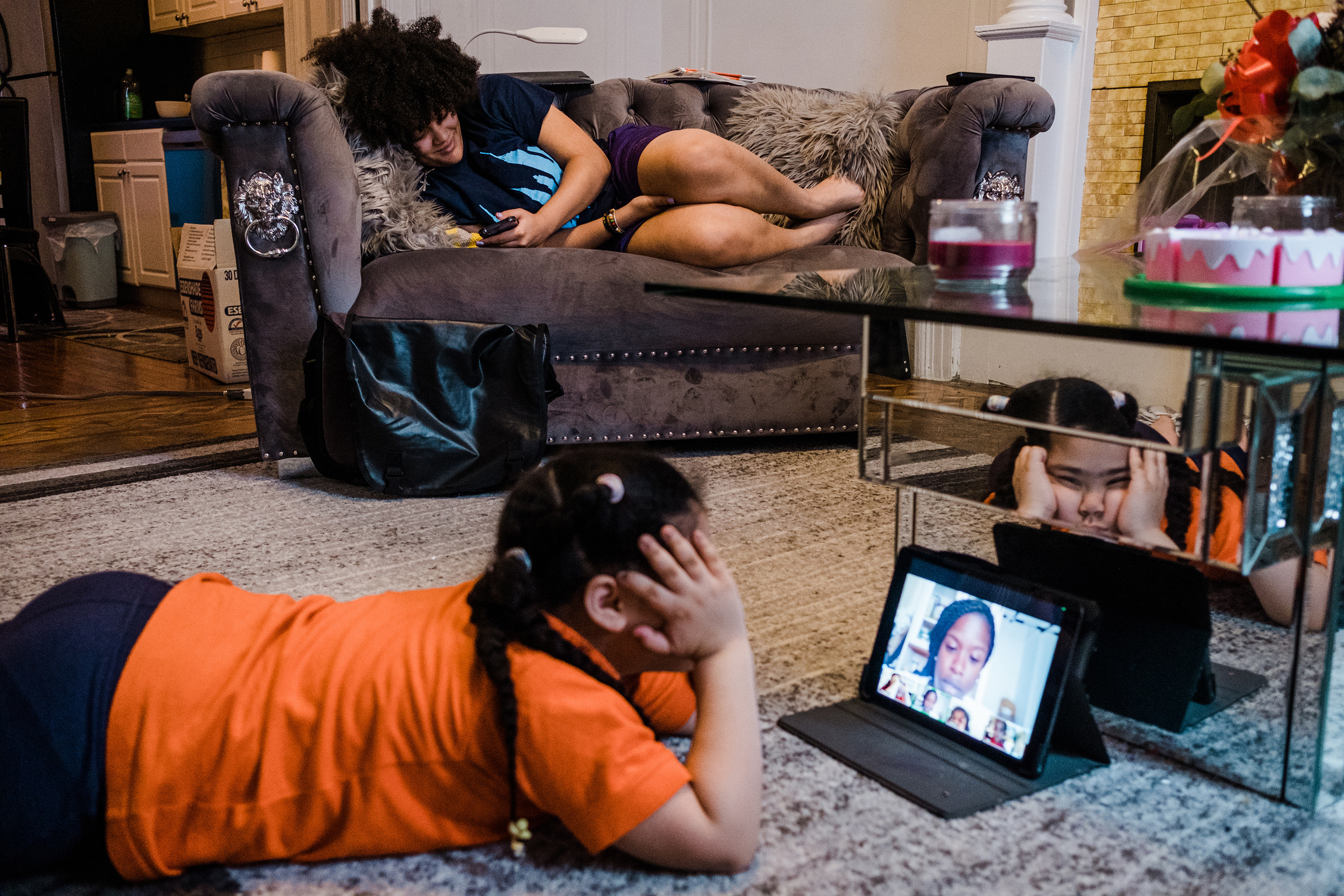 A girl lies on the ground looking at an iPad screen while her sister lies on the couch behind her