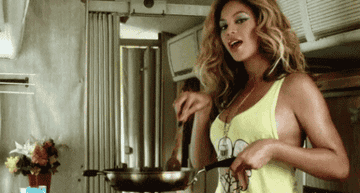 Beyonce stirring food in a pan as she has face full of glamorous makeup and a tank top