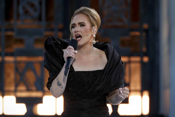 Adele singing onstage in a long gown with a dramatic neckline and drop earrings in the shape of Saturn