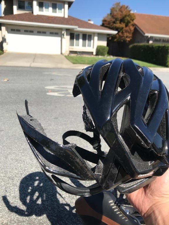 Hand holds a broken bike helmet in a street with houses in the background, highlighting helmet damage and potential accident