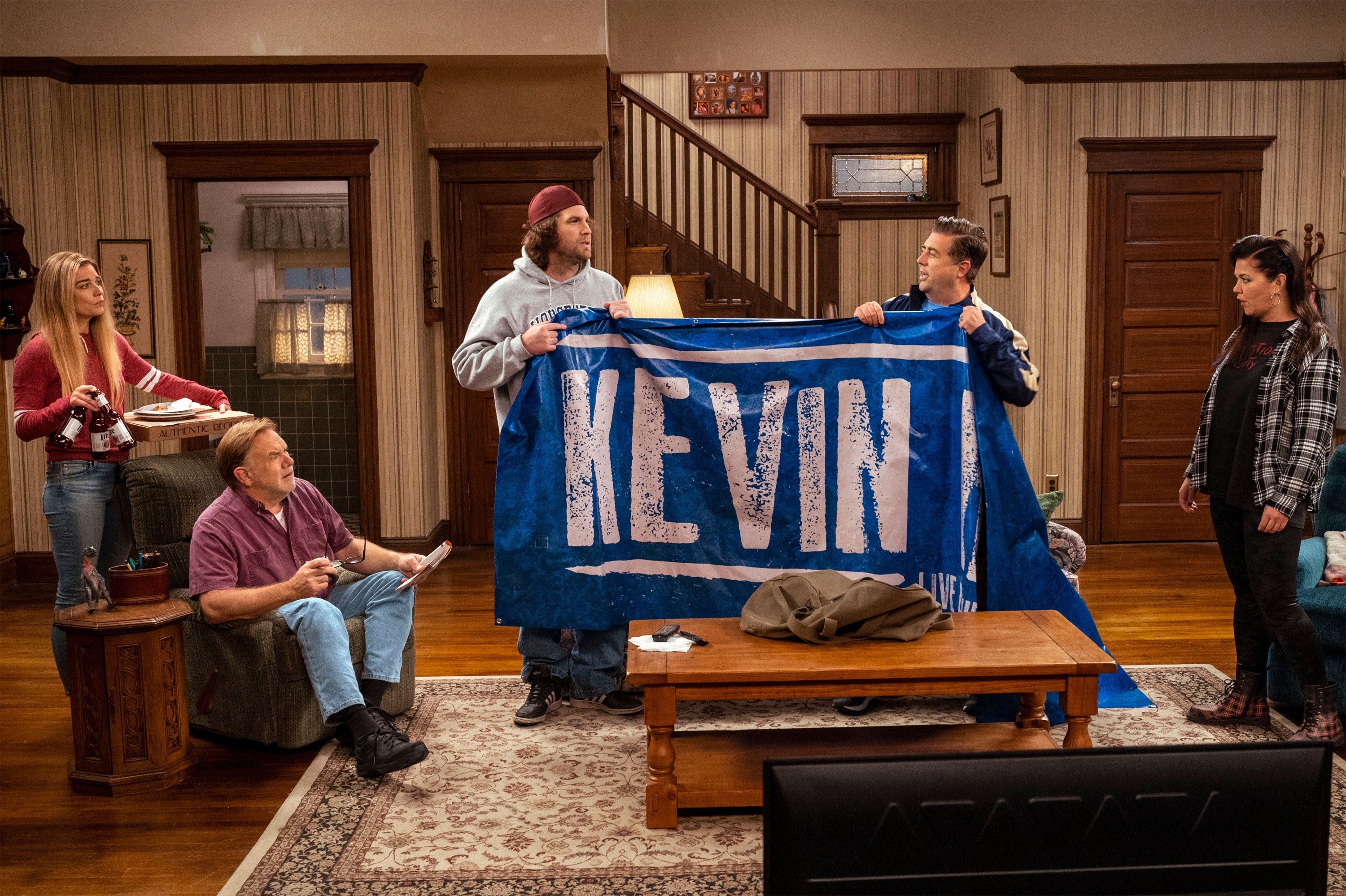 In the sitcom living room, kevin&#x27;s friends hold up a giant blanket reading &quot;Kevin&quot;