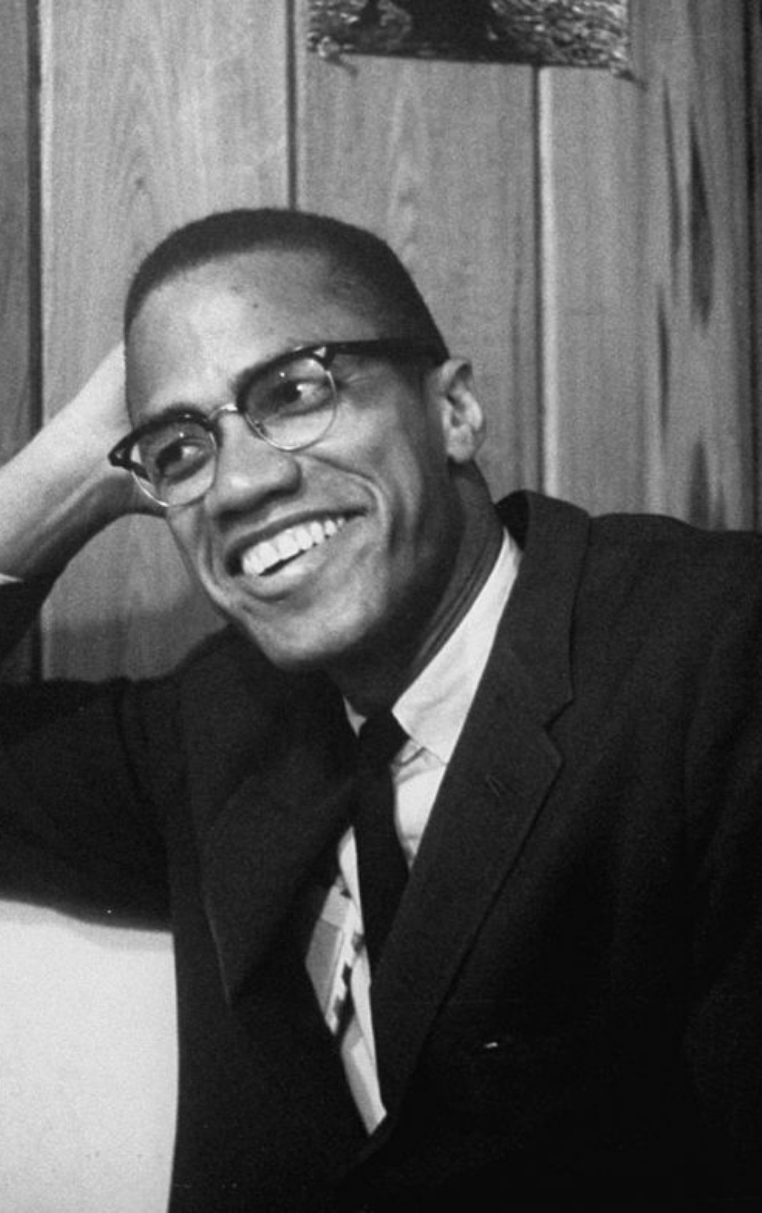 Malcolm X smiling while leaning his arm on the side of a booth