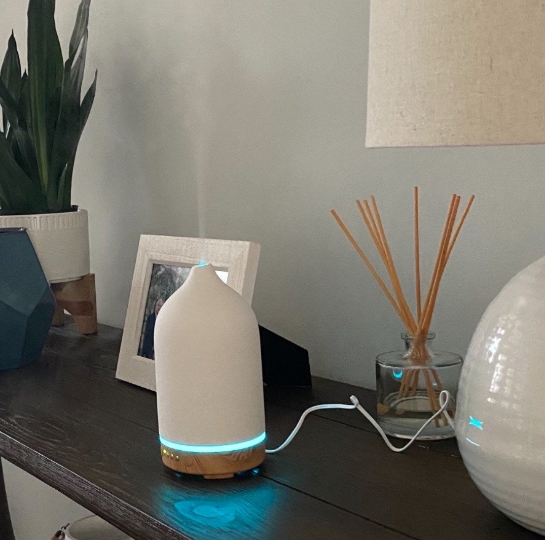 a reviewer's 100ml diffuser sitting on a table beside incense and picture frames. The diffuser has a wooden base and a cream-colored top.