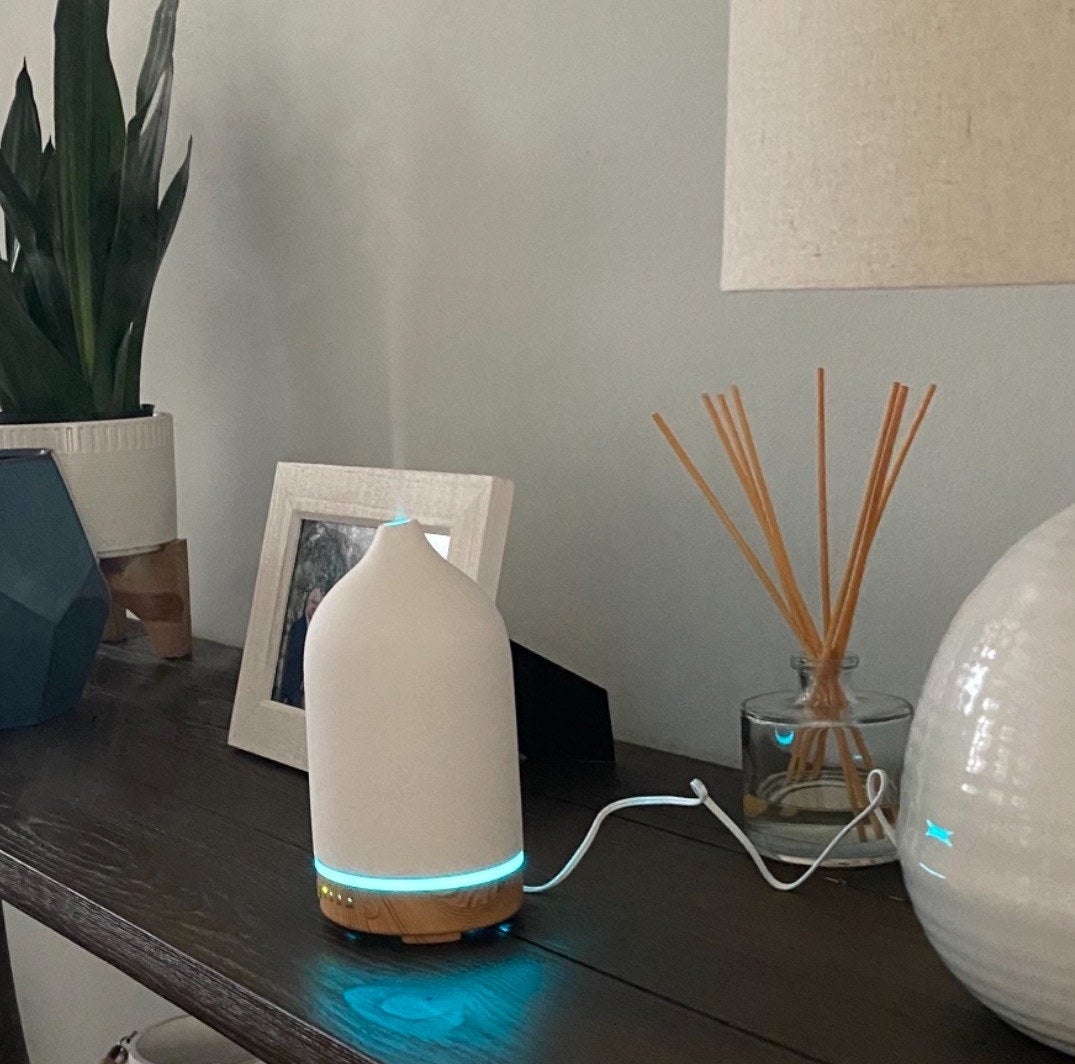 a reviewer&#x27;s 100ml diffuser sitting on a table beside incense and picture frames. The diffuser has a wooden base and a cream-colored top.