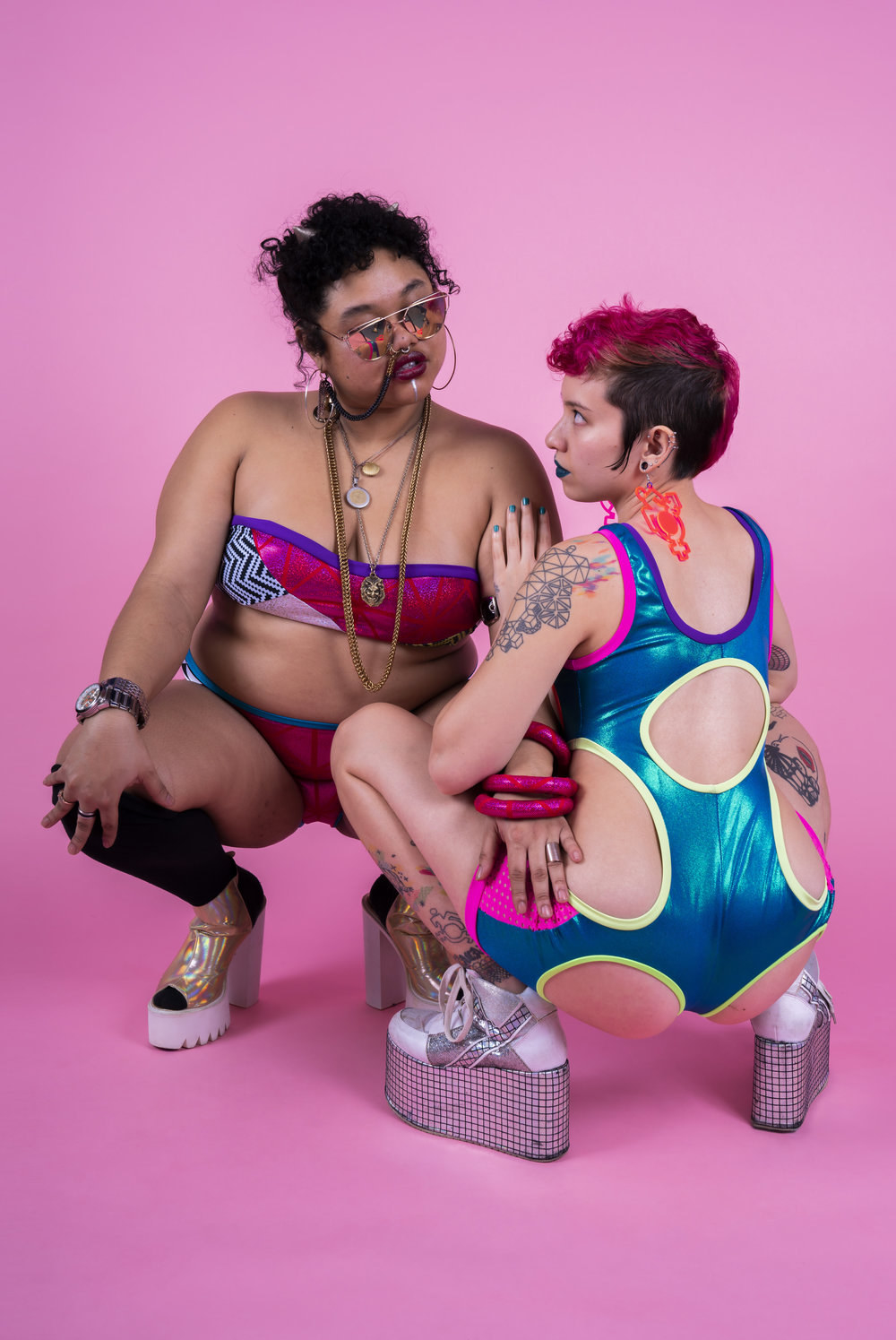 Two humans kneeling together, one in a brightly colored two piece bandeau set with jewelry, another in a shiny blue one-piece with multiple cut-outs, both wearing platform heels