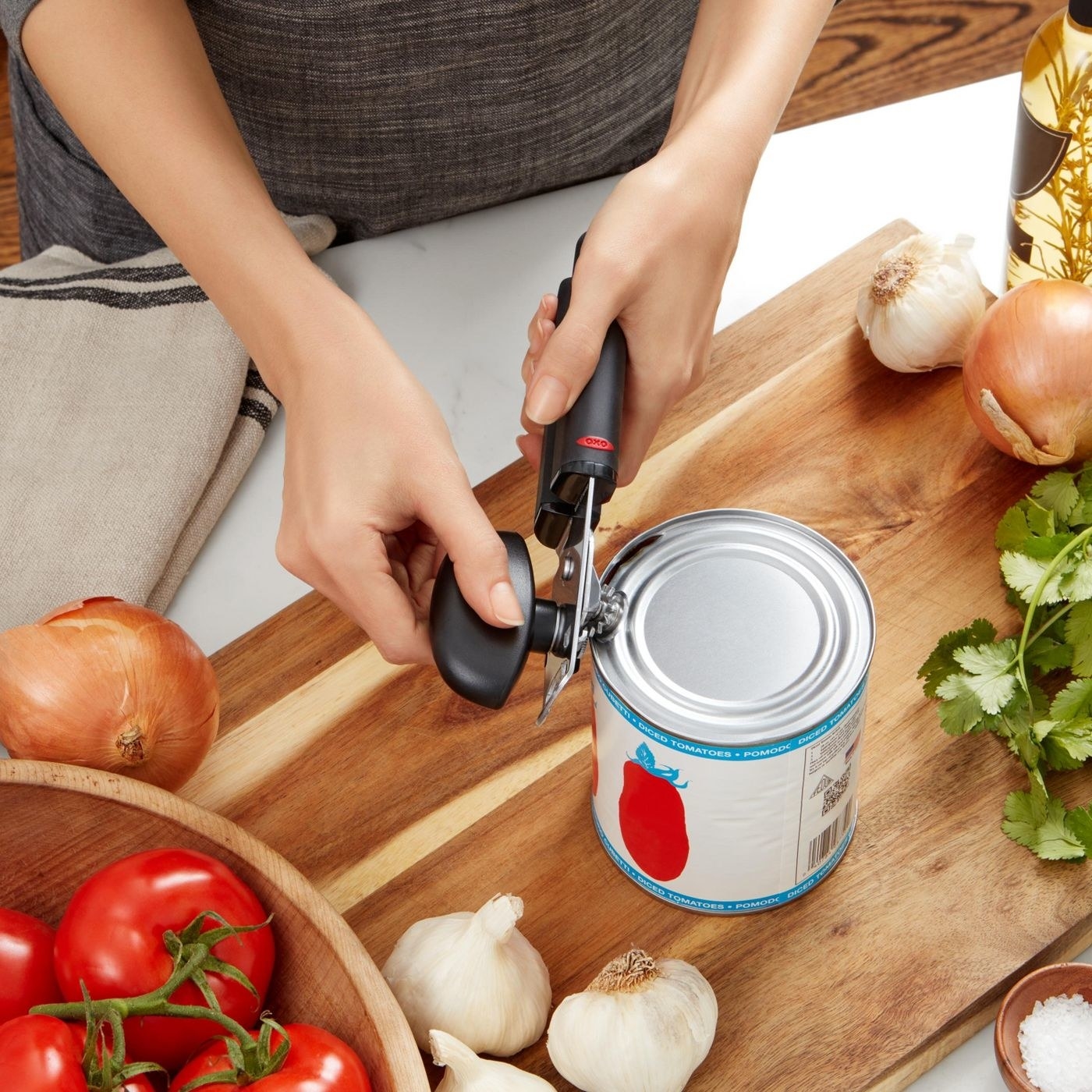 Model using can opener on tomato can surrounded by onions, garlic, tomatoes and herbs