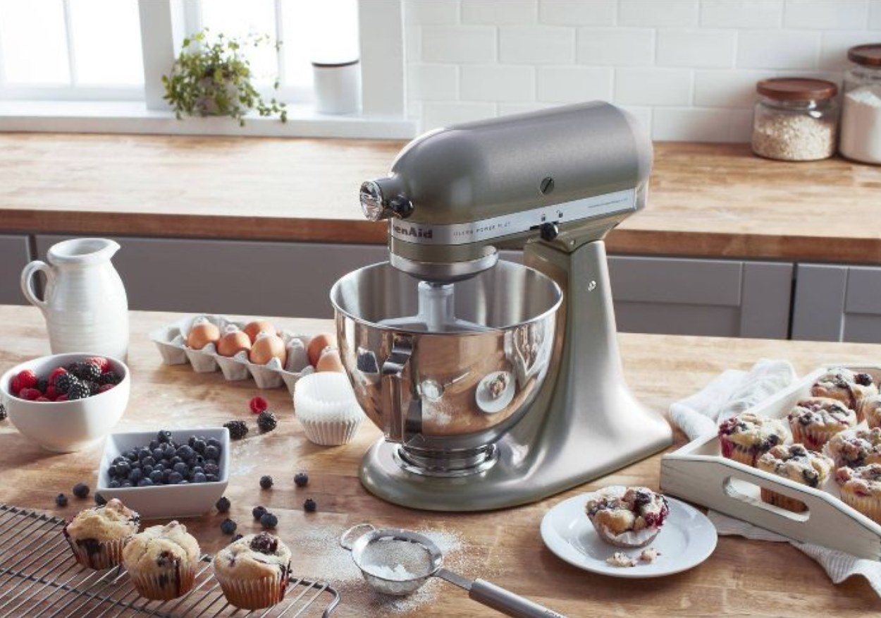 A gray stand mixer with paddle attachment and silver mixing bowl surrounded by baked goods