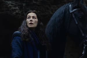 Moiraine looks over her shoulder as she stands beside a black horse.