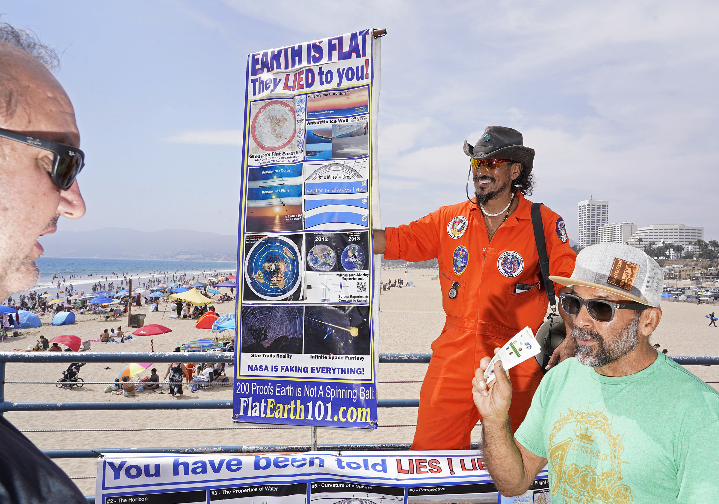 Three men in sunglasses, one in an astronaut jumpsuit holding a sign saying &quot;the earth is flat they lied to you&quot;