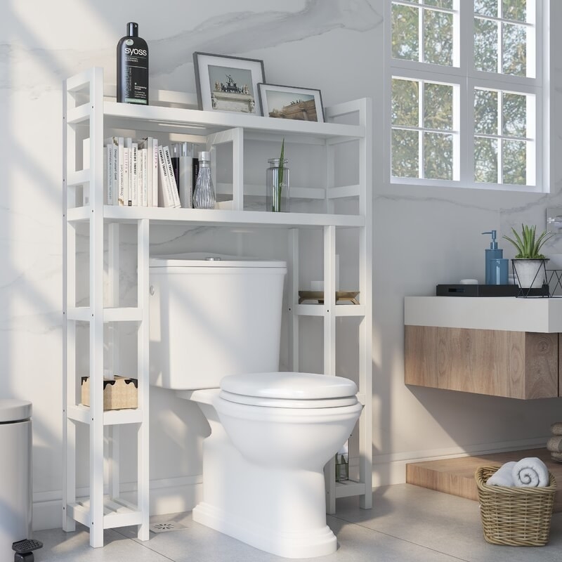 A white over-the-toilet cabinet with various books and decor on the shelves
