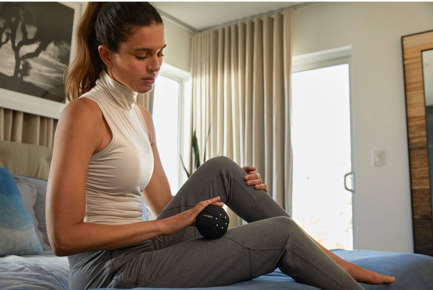 A woman leans on her bed and uses a mini vibrating massage ball on her thigh.