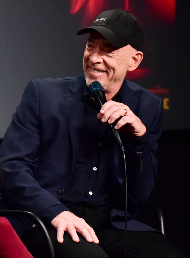 A smiling J.K. sitting, wearing a cap, and holding a microphone