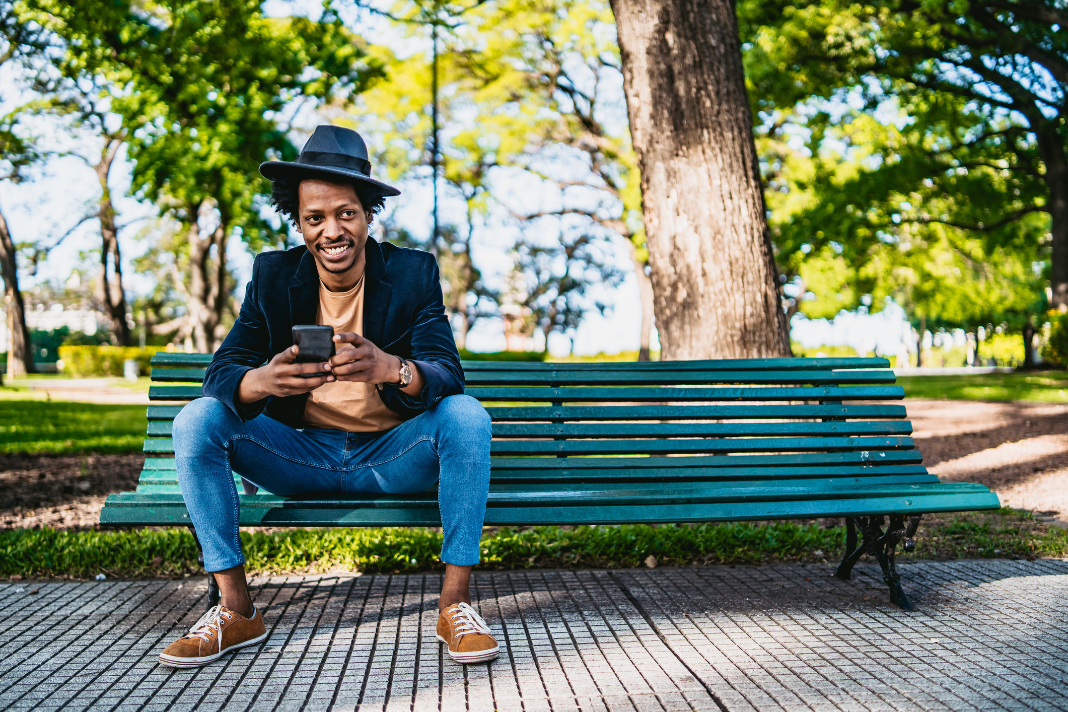 Smiling man wearing a hit and sitting on a park bench holding a cellphone