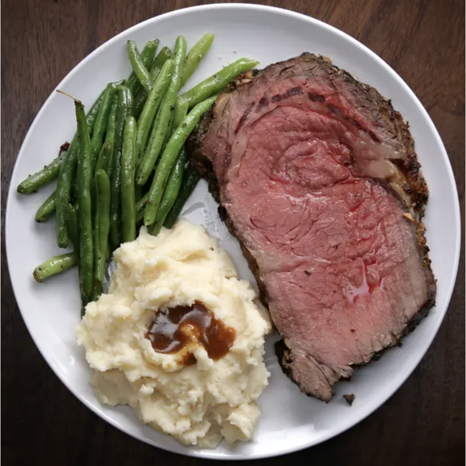 An overhead shot of a white plate with a juicy cut of prime rib alongside mashed potatoes with gravy and cooked green beans