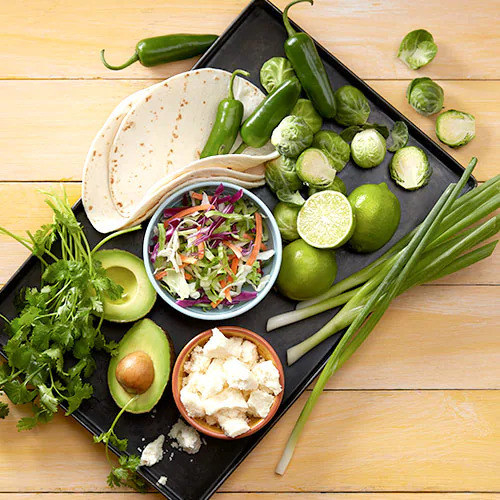 serving platter with tortillas, Brussels sprouts, avocado, sliced limes, and cheese