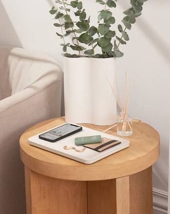 a white catch tray charging station sitting on a side table with a phone, wallet, and keychain on top.
