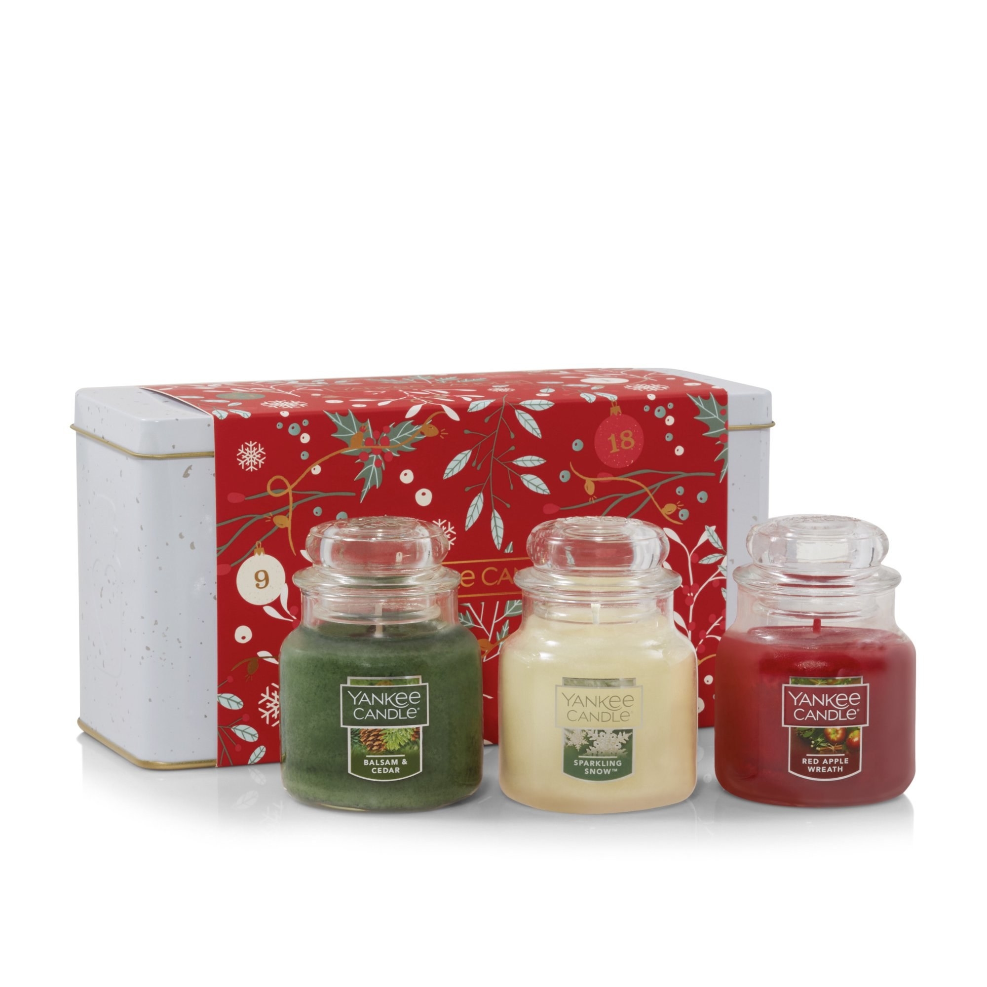 Three Yankee Candles displayed in front of the metal container they are packaged in: Balsam &amp;amp; Cedar, Sparkling Snow, and Red Apple Wreath