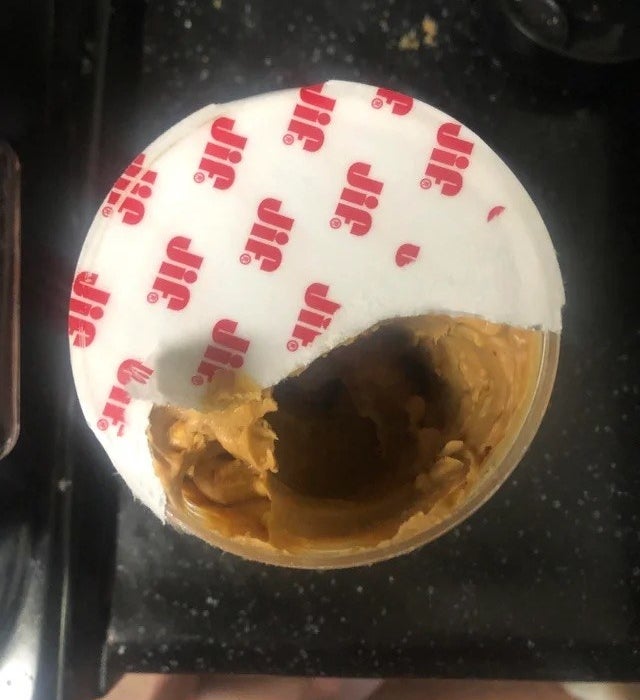 A jar of peanut butter where the paper liner is still mostly on and someone only ripped part of it off