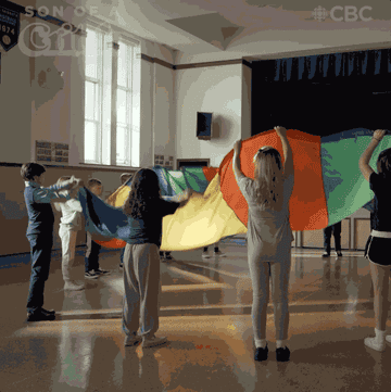 An animated GIF a class in a circle using a multicoloured parachute in the gym.