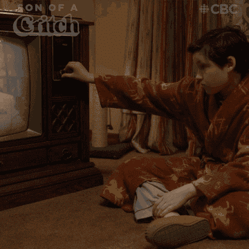 A boy sitting in front of an old tv in a robe turning the dial.