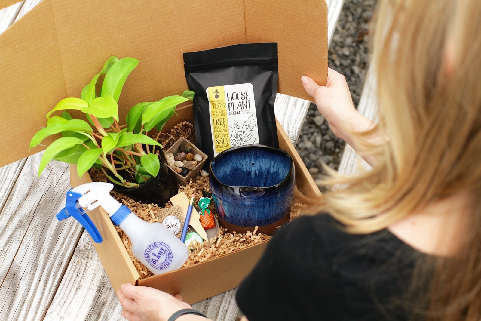 A person opening up a box with a plant, water bottle, pot, and other plant-related products inside