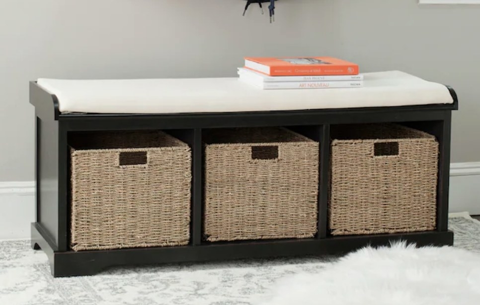 A black wooden storage bench with a cream cushion atop and 3 cubbies with wicker baskets