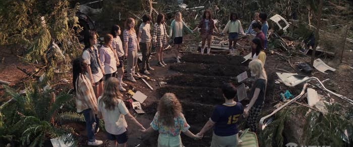 A bunch of girls and two boys hold each other's hands and form a circle around the grave of some people