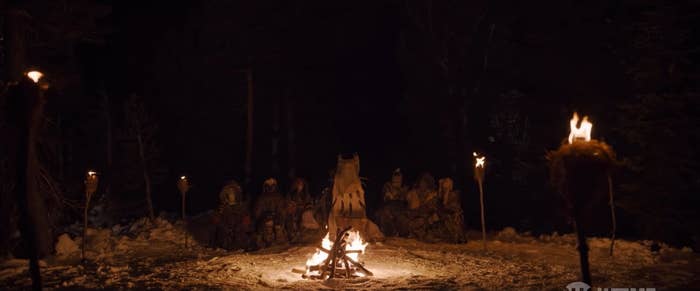 A group of girls dressed in animal skin, sit around a bonfire