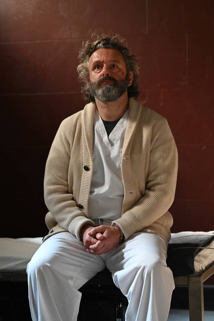Sheen sits on a bed clasping his hands with a cut on his face as he acts in a scene from a play