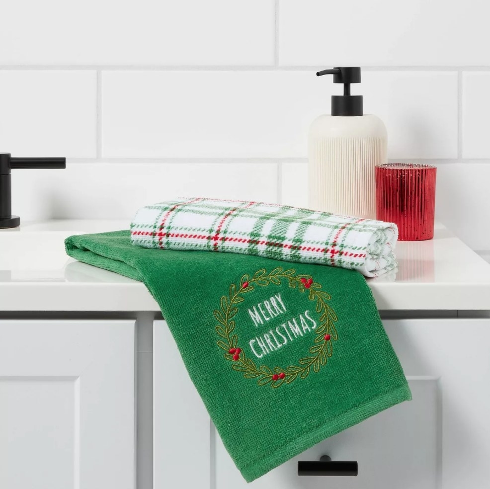Green towel that says merry christmas with wreath around it, plaid green white and red towel folded on top of it, both on white countertop