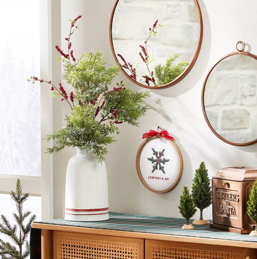 Hoop art with green snowflake that says &quot;comfort &amp;amp; joy&quot; hanging on wall with round mirrors above mantle, next to holiday plant in white vvase