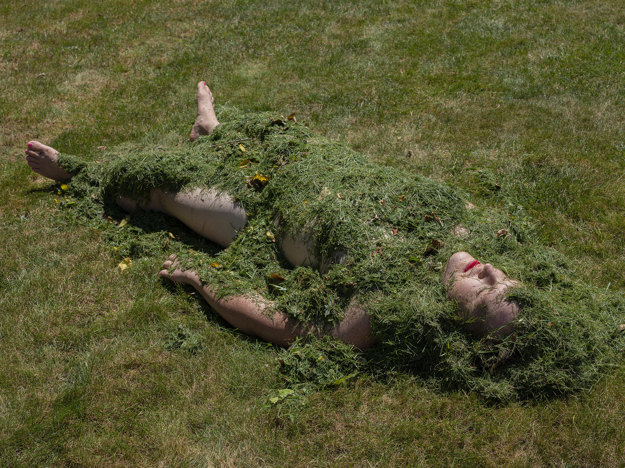 A person lies on the ground covered in grass clippings with red lipstick 