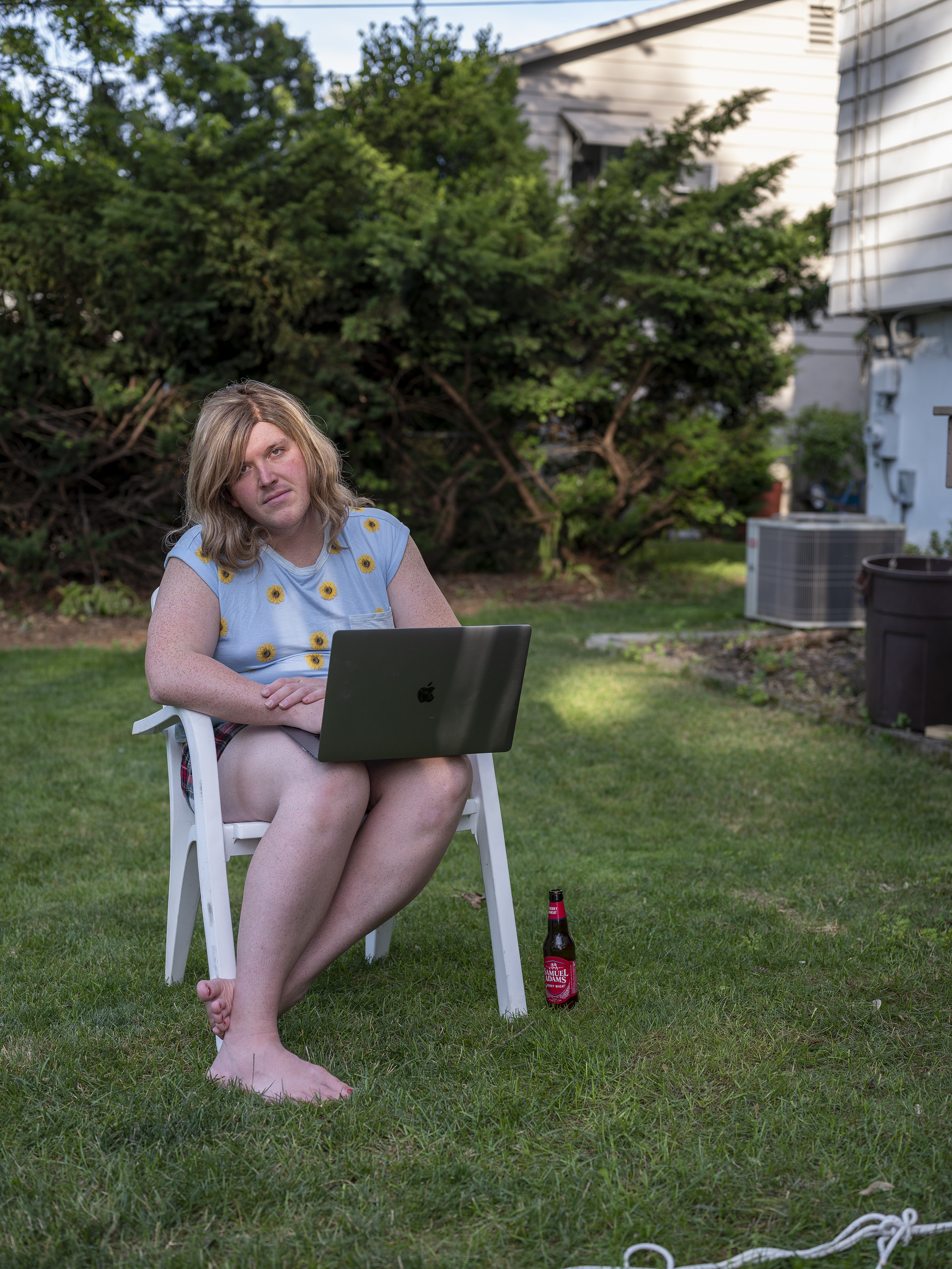 a person in a wig sits in a chair on their lawn with a laptop computer and drinking a beer